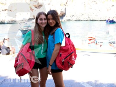 Due studentesse sorridenti con zaini posano davanti a un porto turistico maltese, tra barche, acqua e costa rocciosa.