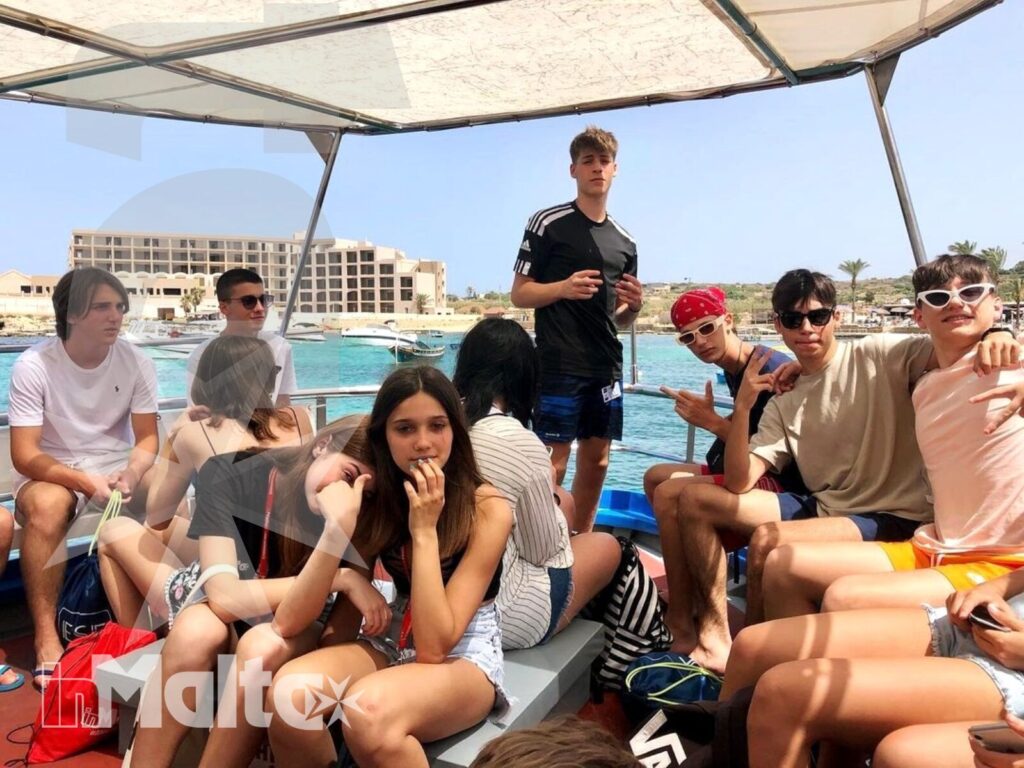 Gruppo numeroso di studenti siede e sta in piedi su una barca, con il porto e la costa di Malta sullo sfondo.