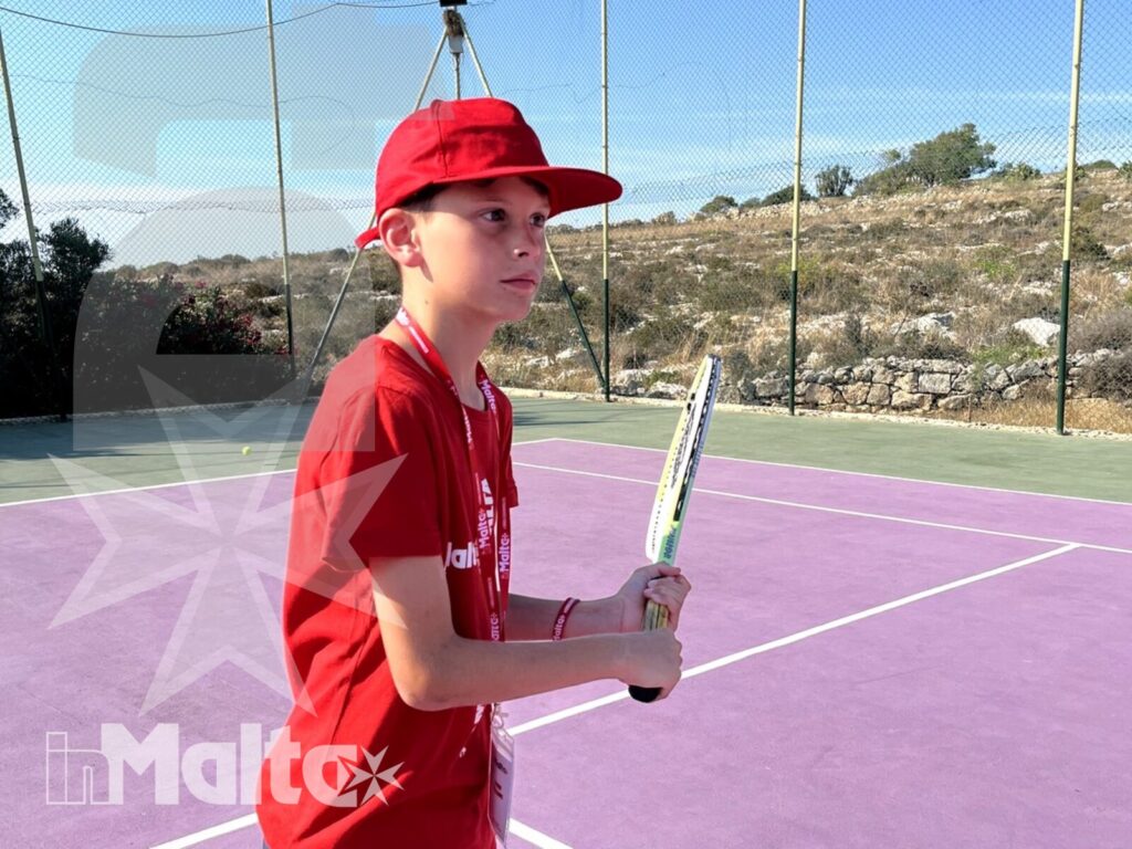 Studente con racchetta e abbigliamento sportivo rosso sul campo da tennis all'aperto del Salini Resort, in un paesaggio mediterraneo di Malta.