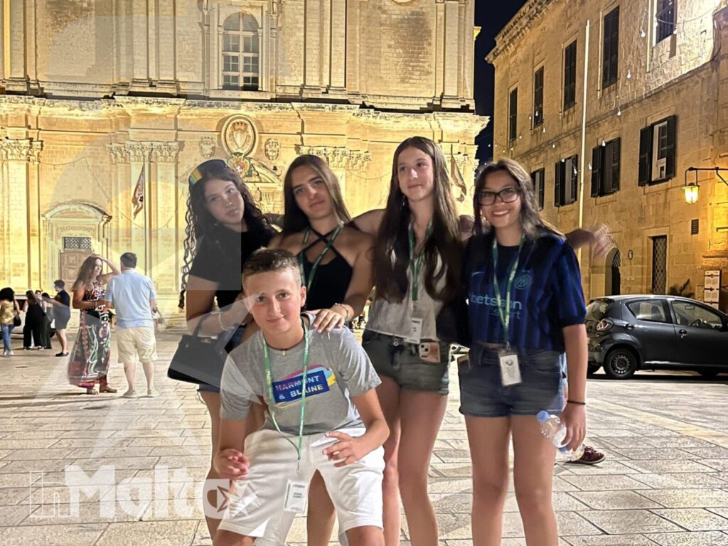 Cinque studenti sorridono in una piazza storica di Mdina, davanti a una chiesa barocca illuminata durante la visita serale.