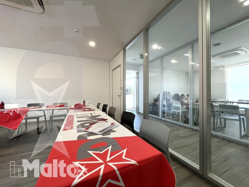 Tavoli apparecchiati con tovaglie rosse e materiali cartacei in una sala interna moderna collegata ad altri ambienti vetrati.