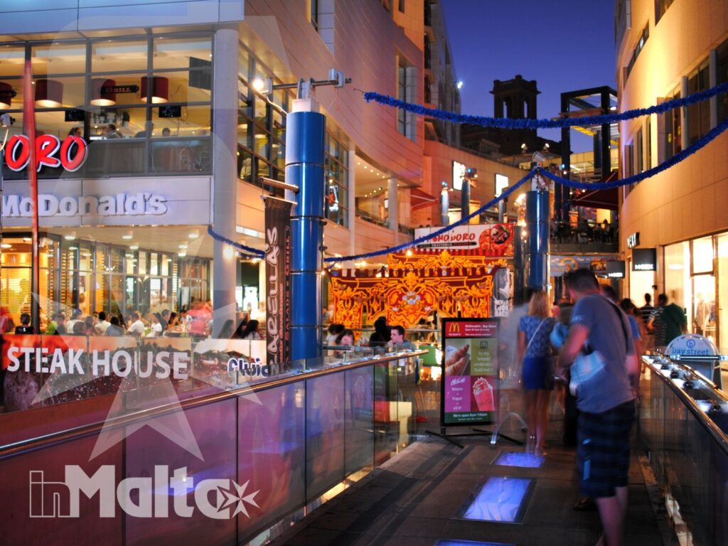 Galleria commerciale e passeggio serale nel centro di Paceville Galleria commerciale all'aperto al crepuscolo, con ristoranti, decorazioni luminose e persone a passeggio nel centro di Paceville.