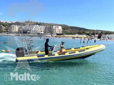 Gommone giallo e nero con due persone naviga davanti a una spiaggia mediterranea, tra acqua turchese, ombrelloni e costa urbanizzata di Malta.
