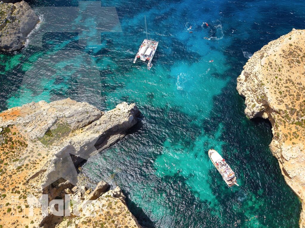 Caletta di Comino vista dall'alto durante l'escursione Caletta turchese di Comino ripresa dall'alto, con rocce chiare, due imbarcazioni e bagnanti nell'acqua limpida.