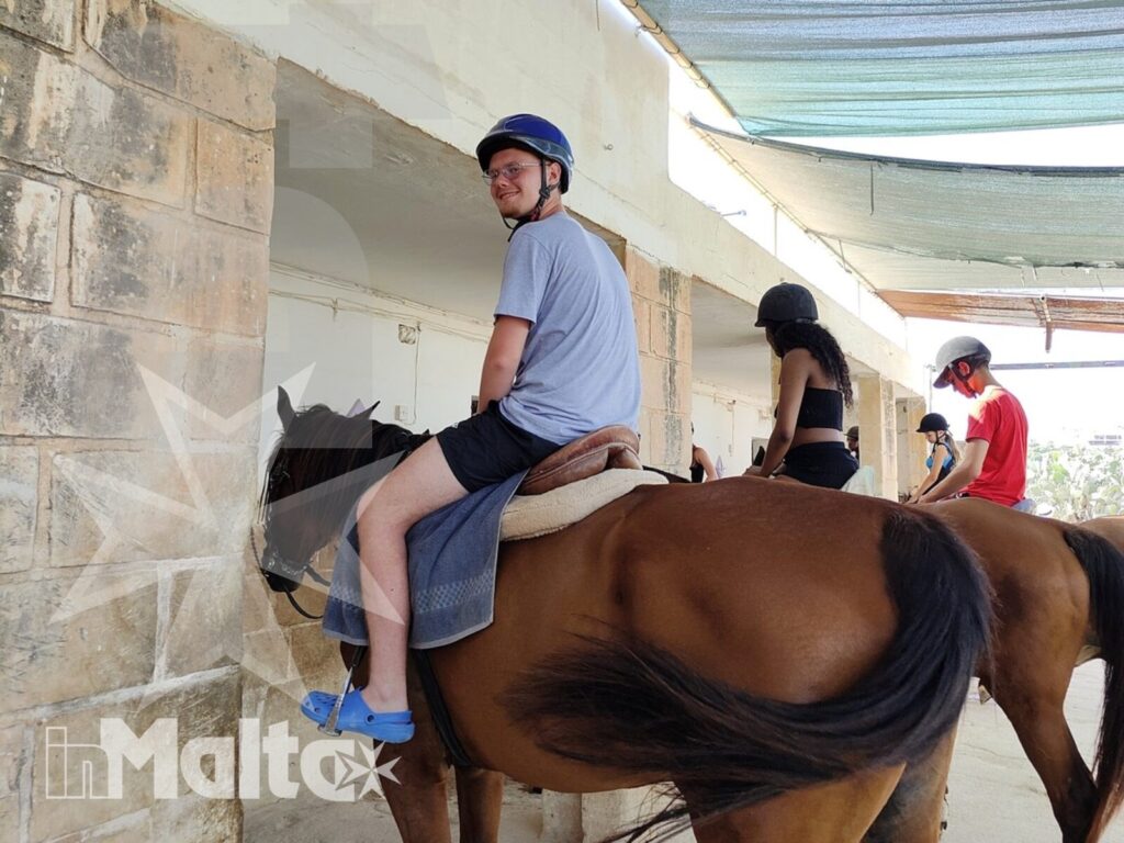 Studente a cavallo sotto una tettoia ombreggiante, seguito da altri partecipanti in fila durante l'attività equestre.
