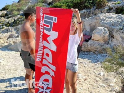 Due studenti mostrano una bandiera del gruppo tra rocce calcaree e vegetazione mediterranea durante un'attività all'aperto a Malta.