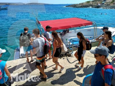 Studenti con zaini salgono su una barca dal tendalino rosso in una baia maltese dalle acque turchesi.