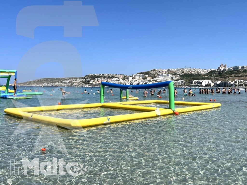 Water Games inMalta tra gonfiabili, mare e costa mediterranea Parco gonfiabile colorato in acque basse e trasparenti, con bagnanti sullo sfondo e una costa mediterranea urbanizzata.