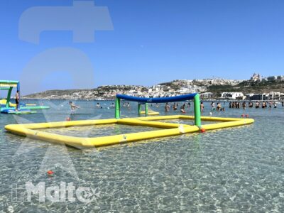 Parco gonfiabile colorato in acque basse e trasparenti, con bagnanti sullo sfondo e una costa mediterranea urbanizzata.