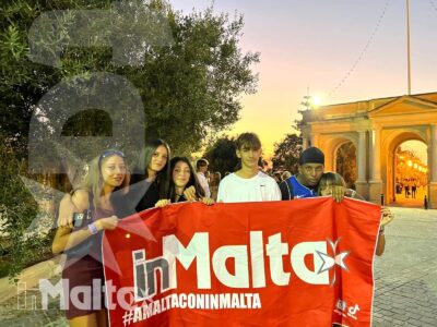Studenti internazionali posano con una bandiera di Malta davanti a un monumento illuminato, durante una visita serale.