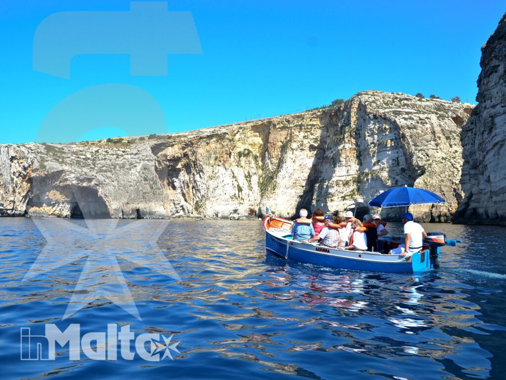 Gita in barca tra le scogliere della Blue Grotto Barca turistica blu e bianca con passeggeri naviga davanti alle scogliere calcaree della Blue Grotto, sotto un cielo sereno.
