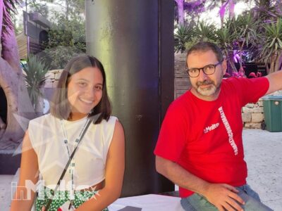 Due persone sorridono sedute davanti al pannello INMALTA, immerse nella vegetazione durante un'attività del gruppo.
