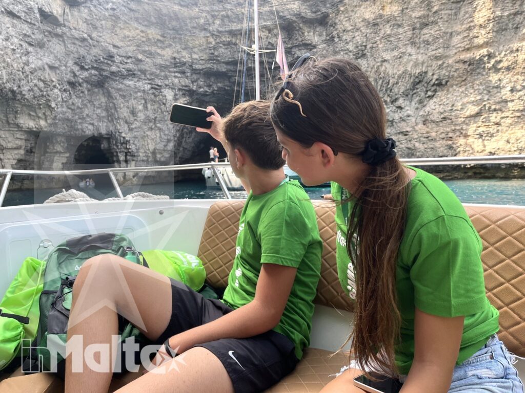 Due studenti osservano una grotta marina dalla barca mentre uno fotografa la costa rocciosa di Malta.