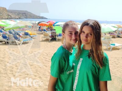 Due studentesse sorridenti siedono sulla spiaggia di Golden Bay, con ombrelloni colorati e mare sullo sfondo in una giornata estiva.