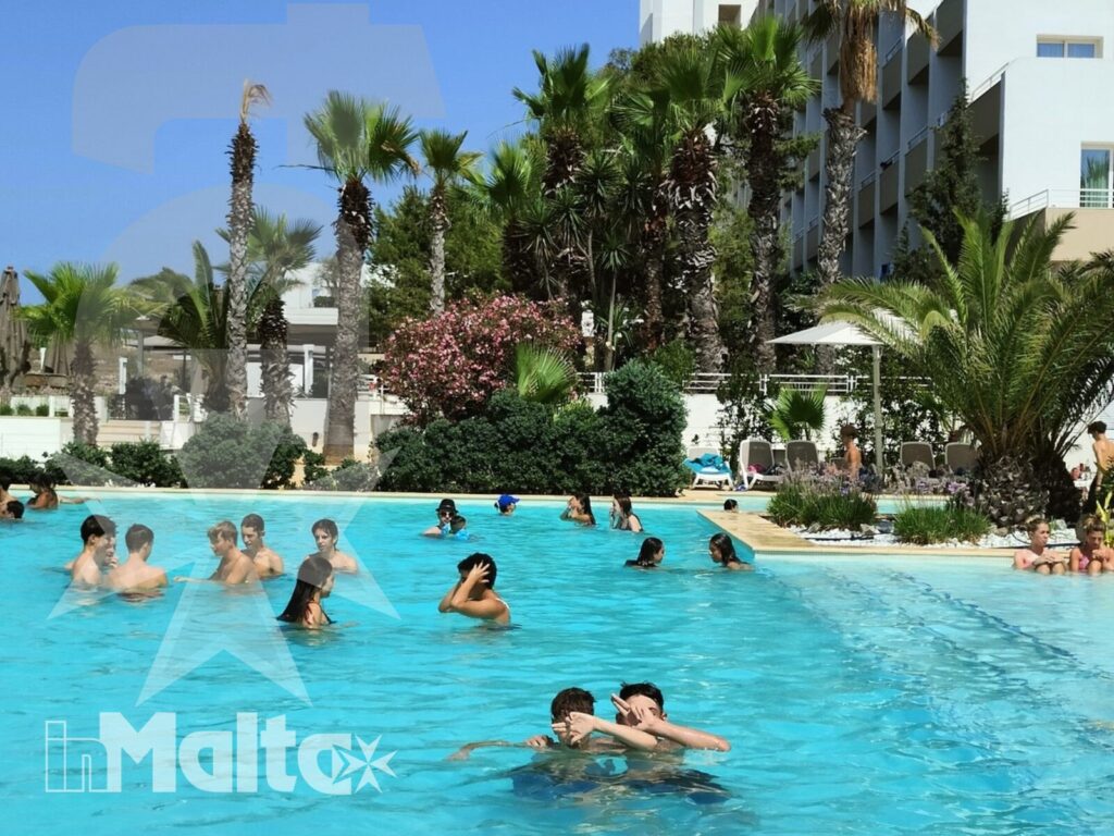 Studenti e ospiti nuotano nella grande piscina turchese del resort, circondati da palme e vegetazione in un'atmosfera estiva.