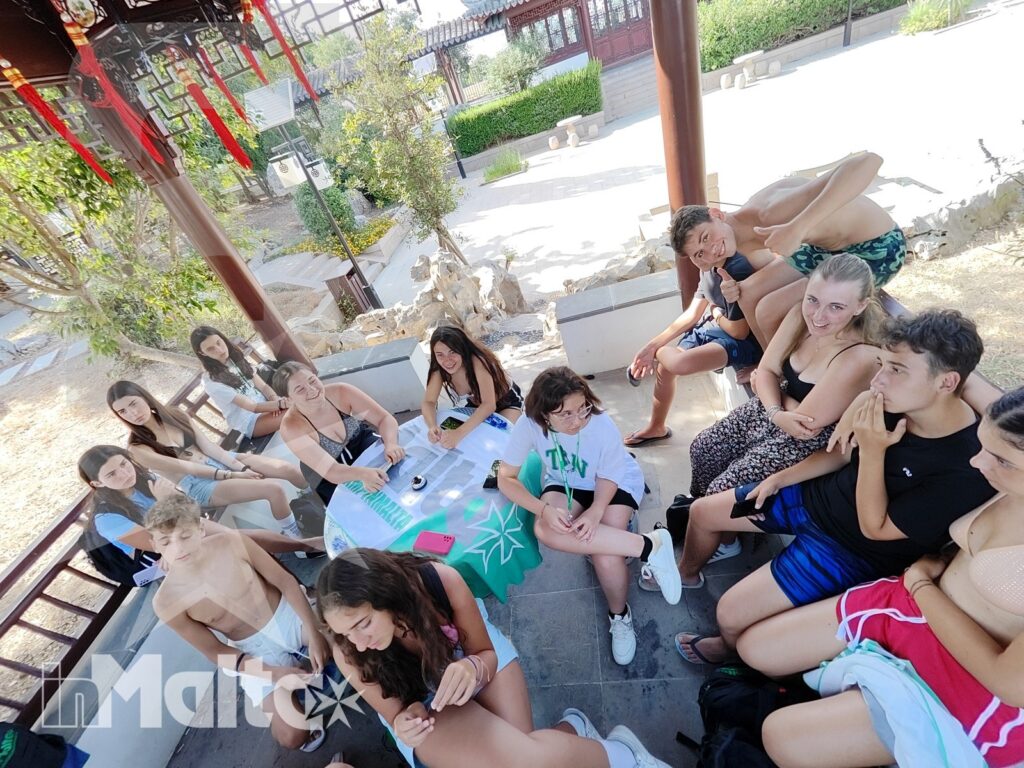 Gruppo di studenti si rilassa e conversa sotto un pergolato tradizionale durante un'attività sociale nei giardini cinesi.