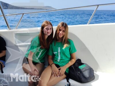 Due studentesse sorridono a bordo di una barca durante un'escursione, con il mare e la costa mediterranea alle spalle.