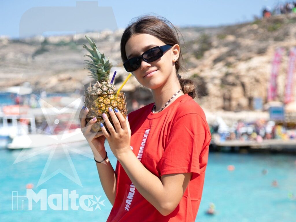 Studentessa inMalta con ananas sulla costa Studentessa con maglietta rossa mostra un ananas davanti al mare turchese durante l'escursione a Malta.