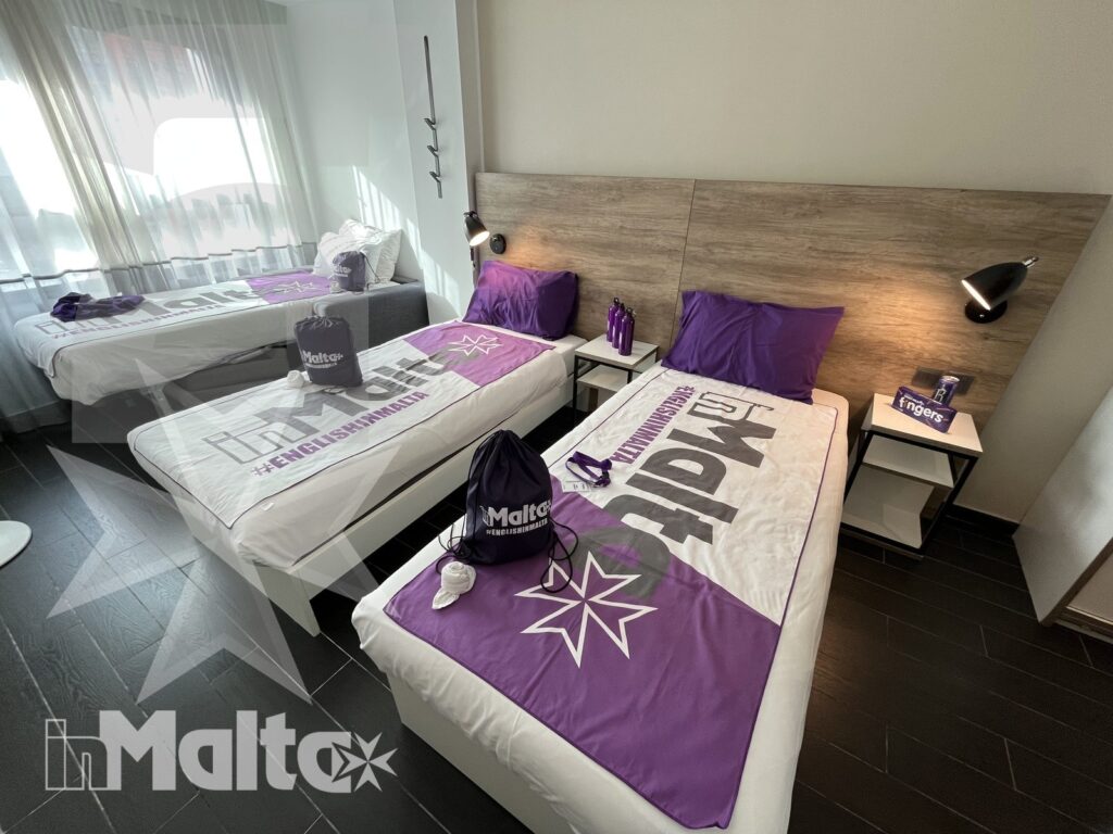 Camera condivisa brandizzata per studenti in soggiorno a Malta Camera condivisa con tre letti singoli, tessili viola e accessori personalizzati, predisposta per studenti e gruppi in soggiorno a Malta.