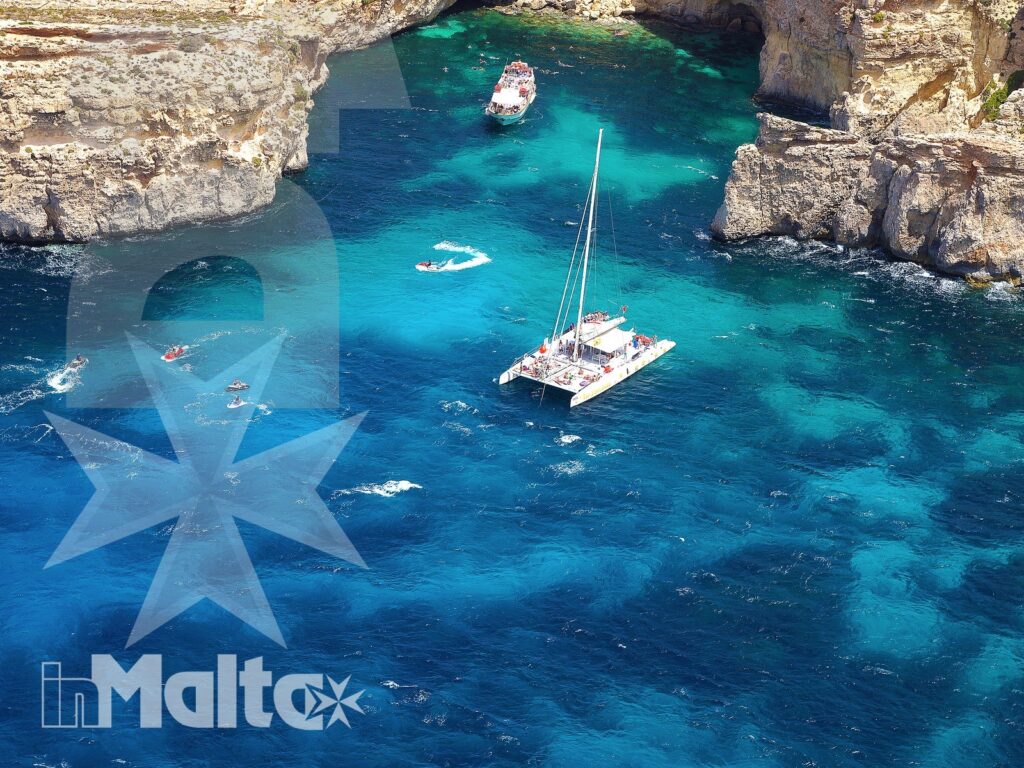 Vista dall'alto della laguna turchese di Comino, con catamarano, piccole barche e bagnanti circondati da scogliere calcaree.