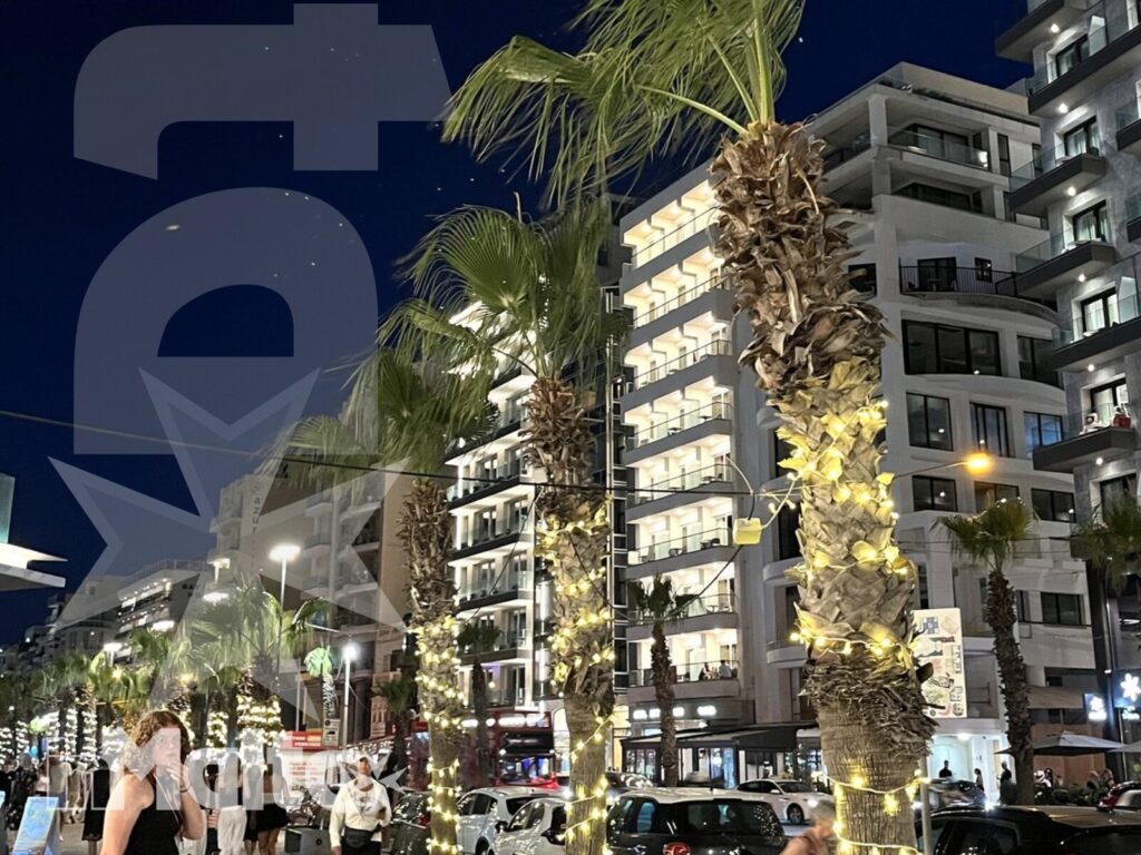 Palme illuminate costeggiano una strada serale con hotel moderni, traffico, pedoni e locali dall'atmosfera mediterranea.