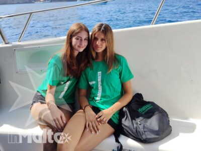 Due studentesse sorridenti siedono a bordo con giubbotti di salvataggio, mentre il mare e la costa rocciosa maltese si aprono sullo sfondo.