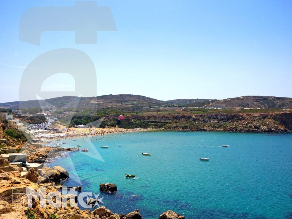 Panorama di Golden Bay tra mare turchese e colline maltesi Veduta panoramica di Golden Bay con acqua turchese, spiaggia affollata, barche e colline terrazzate sulla costa maltese.