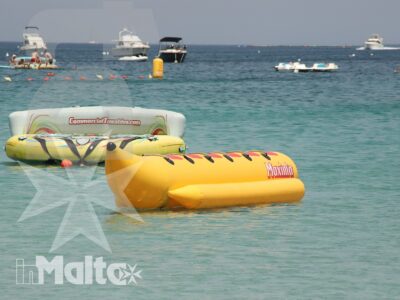 Banana boat e zattera gonfiabile galleggiano in primo piano, mentre barche e bagnanti animano l'area nautica sullo sfondo.