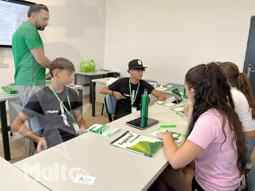 Studenti collaborano in aula durante un'attività guidata Giovani partecipanti discutono su brochure e cartelline attorno a un tavolo, mentre un adulto supervisiona l'attività in aula.