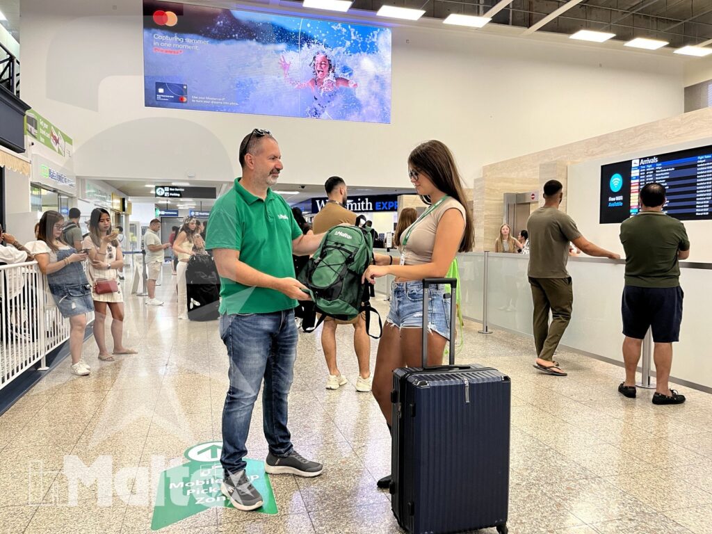 Accoglienza individuale di una studentessa all'aeroporto di Malta Accompagnatore con maglia verde accanto a una studentessa con zaino e bagagli nell'area arrivi dell'aeroporto di Malta.