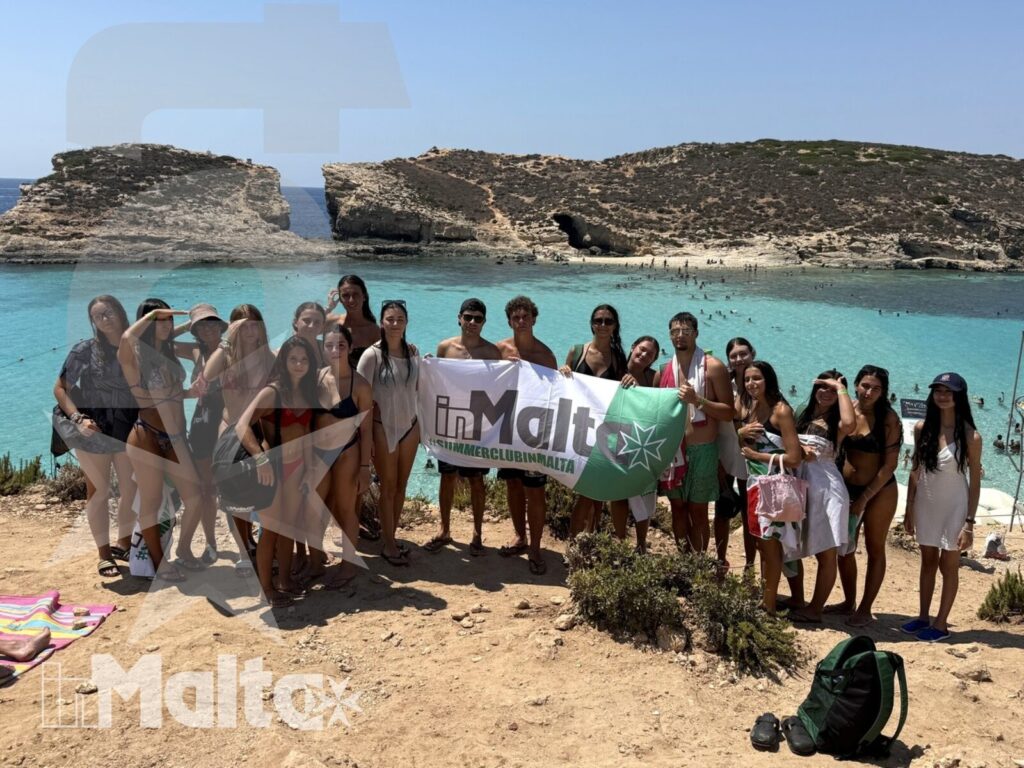 Gruppo internazionale di studenti posa con uno striscione davanti a una baia turchese di Malta, tra scogliere e attrezzatura da spiaggia.