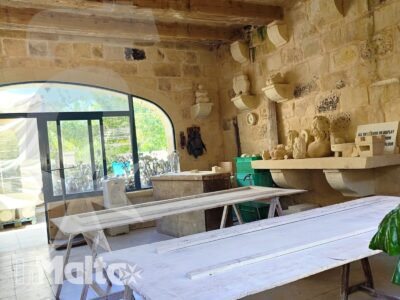 Laboratorio luminoso con pareti in pietra calcarea, travi lignee, tavoli da lavoro e oggetti artigianali in un edificio storico maltese.