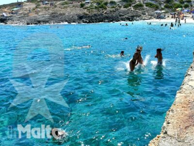Bagnanti nuotano e camminano nella piscina naturale di Hondoq Bay, tra acqua turchese e rocce della costa di Gozo.