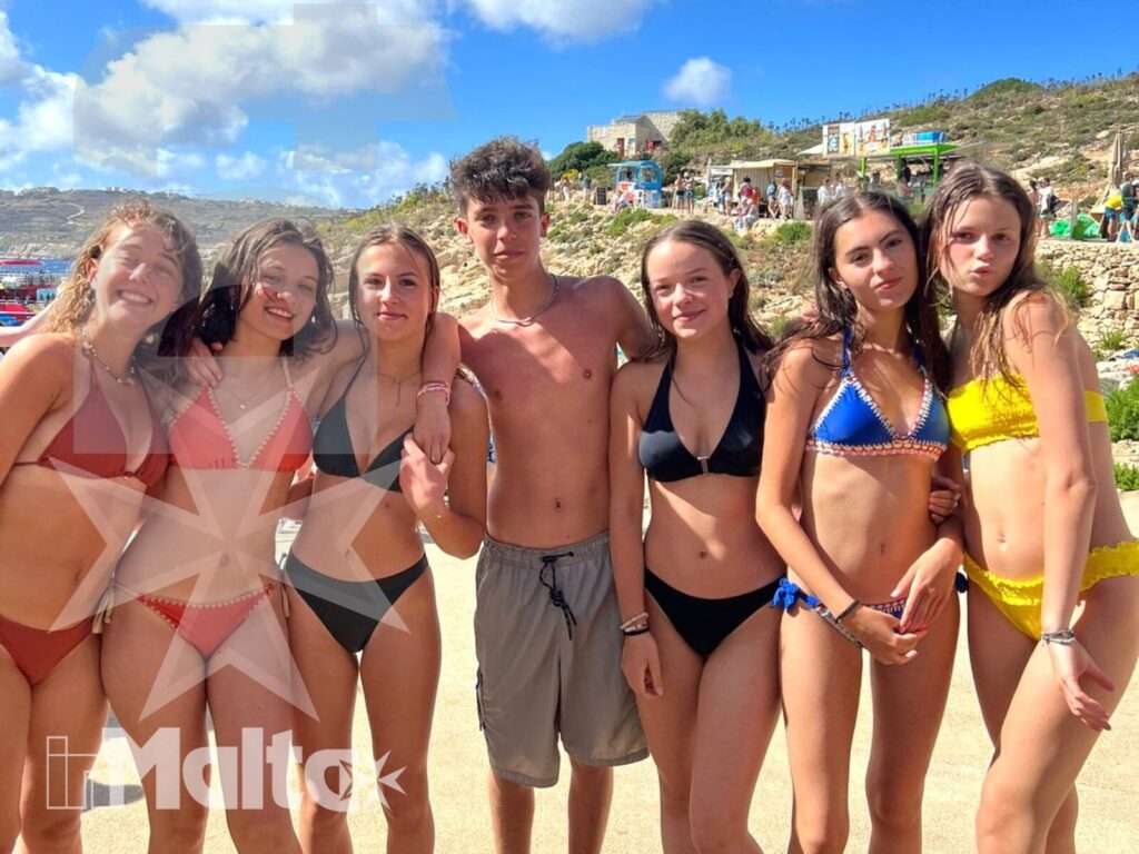 Partecipanti sulla spiaggia durante l'escursione a Malta Gruppo di sette studenti posa sulla spiaggia mediterranea, con costumi colorati e scogliera sullo sfondo.