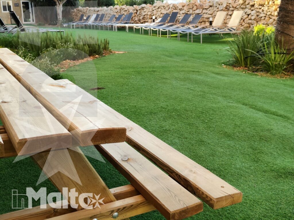 Tavoli da picnic in legno precedono il prato con piscina e lettini, in un'area esterna mediterranea dedicata al relax.