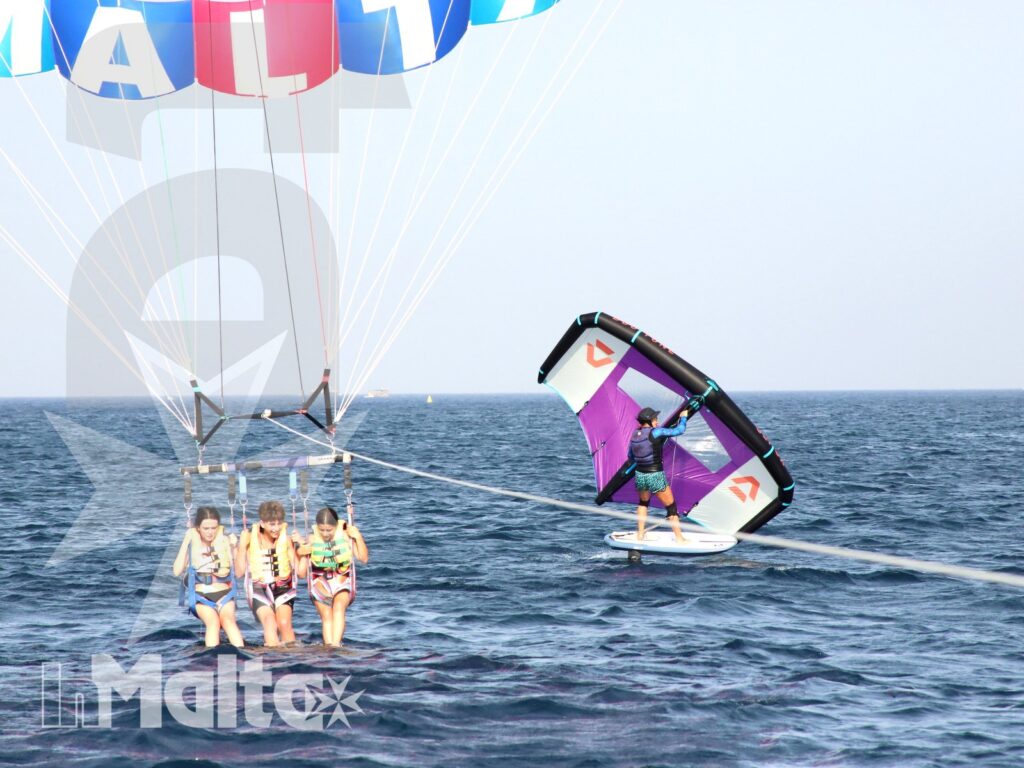 Ragazzi in parasailing sul mare di Malta con un windsurf sullo sfondo durante una giornata di attività acquatiche