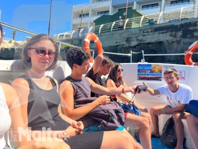 Studenti seduti su una barca ascoltano una guida durante un'escursione in ambiente portuale, con edifici costieri sullo sfondo.