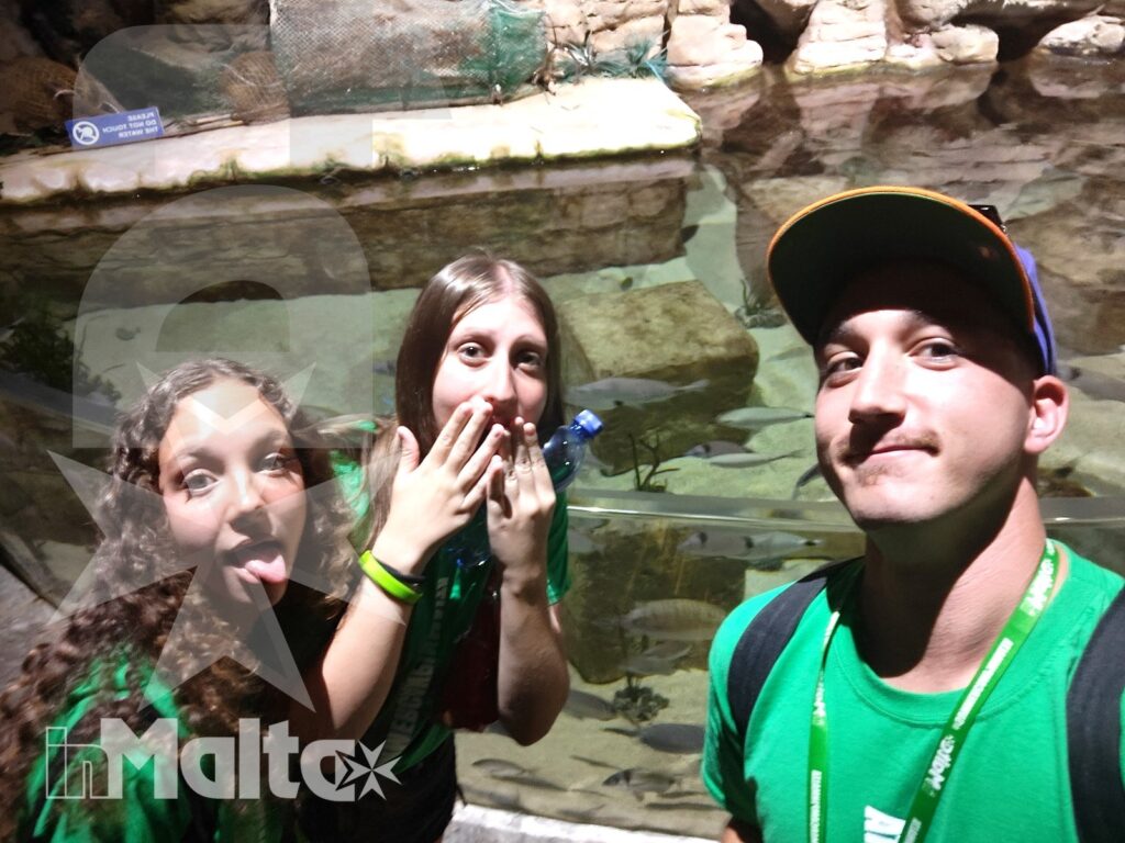 Tre studenti sorridono davanti a una vasca rocciosa durante una visita al Malta National Aquarium, ricordo di una vacanza studio.