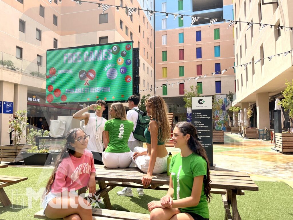 Gruppo di studenti riposa in un moderno cortile con sedute in legno, area verde e grande schermo promozionale.