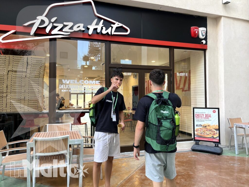 Due studenti con zaini sono davanti all'ingresso di Pizza Hut, tra menu promozionale e tavoli esterni.