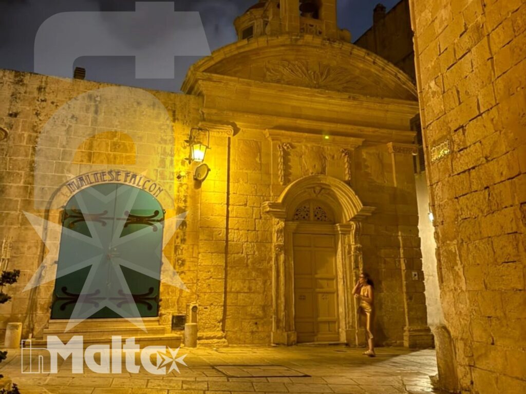 Facciata di una chiesa barocca illuminata nella notte maltese, affacciata su una strada storica in pietra.