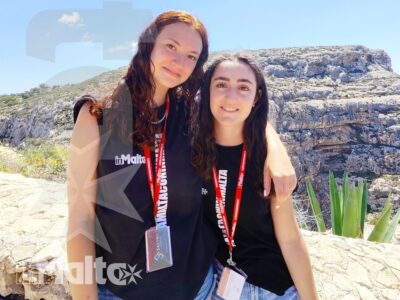 Due studentesse sorridenti e abbracciate durante un'escursione a Malta, con pass identificativi e costa rocciosa mediterranea sullo sfondo.