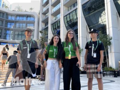 Quattro studenti con badge e zaini posano in un'area pedonale davanti a un moderno complesso urbano durante un'attività inMalta.