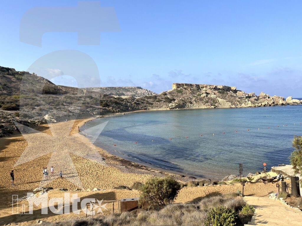 Baia di Malta con spiaggia dorata vista dall'alto Una baia semicircolare con spiaggia dorata, mare calmo e persone in lontananza si apre sotto la vista panoramica della costa maltese.