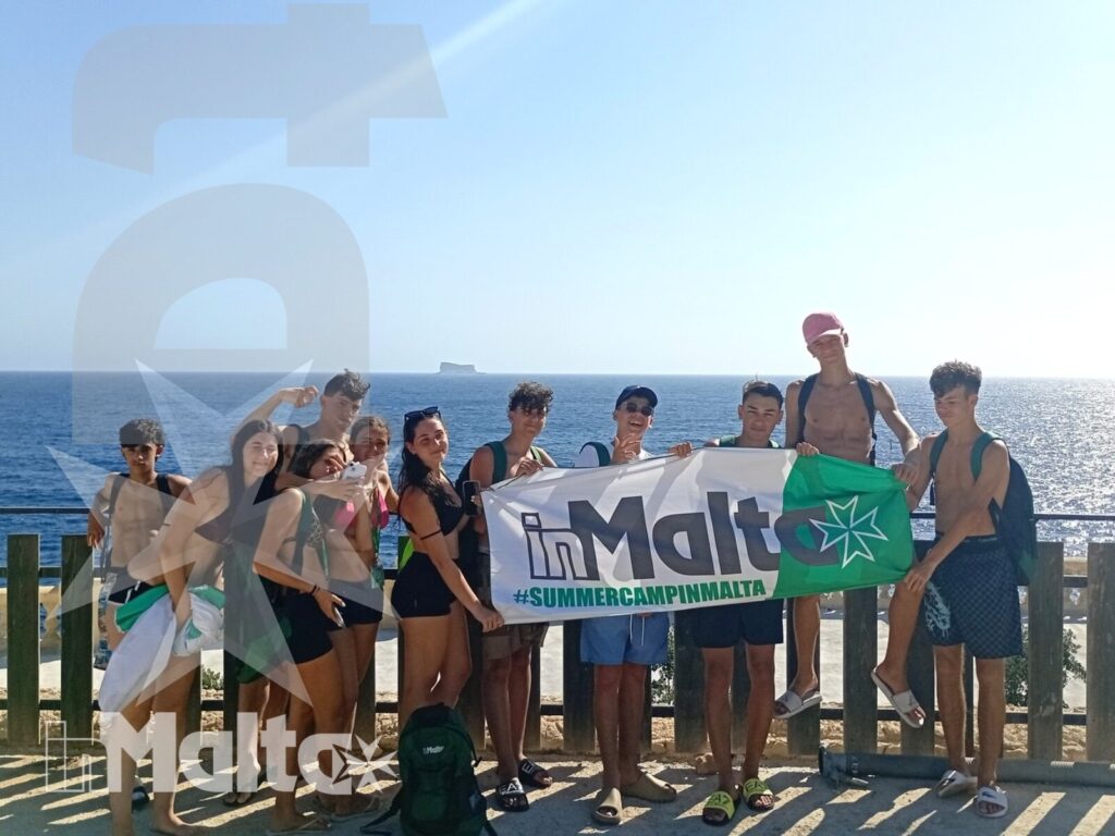 Studenti internazionali posano con uno striscione davanti al mare, indossando abbigliamento estivo durante un soggiorno a Malta.