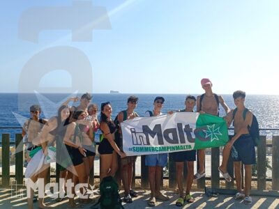 Studenti internazionali posano con uno striscione davanti al mare, indossando abbigliamento estivo durante un soggiorno a Malta.