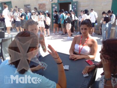 Studentesse internazionali giocano a carte attorno a un tavolo, mentre altri partecipanti socializzano sullo sfondo.