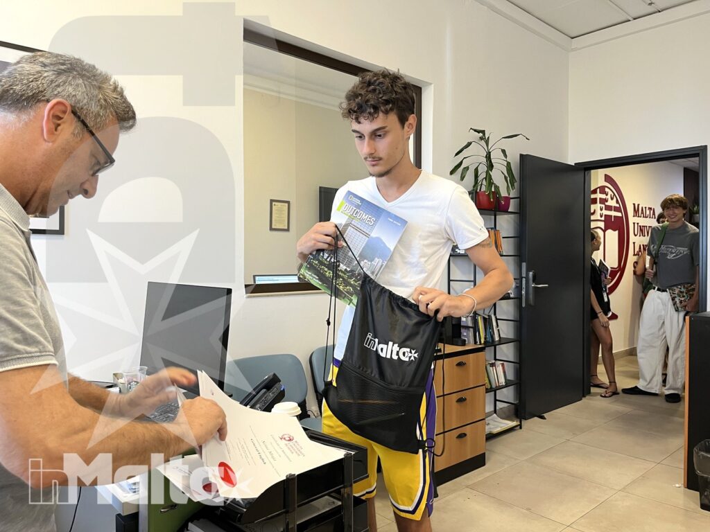 Consegna dei materiali agli studenti durante l'accoglienza Uno studente riceve materiali e organizza una borsa alla reception, assistito da un adulto mentre altri partecipanti attendono.