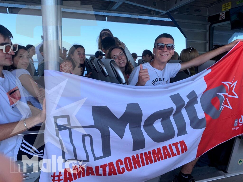 Gruppo numeroso di studenti sorride mostrando una bandiera durante il trasferimento in traghetto verso Malta, in un momento conviviale di viaggio.