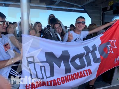 Gruppo numeroso di studenti sorride mostrando una bandiera durante il trasferimento in traghetto verso Malta, in un momento conviviale di viaggio.